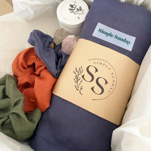 mothers day box consisting of a ink coloured, 100% linen australian made beach bag, 3 XL linen scunchies, ink, rust and olive coloured. White tin candle and rose and clear quartz chunks in white tissue paper.
