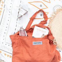 Load image into Gallery viewer, Rust coloured 100% linen, Australian Made beach bag laying down on a towel with water bottle, book and cane hat
