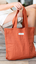 Load image into Gallery viewer, rust coloured 100% linen Australian made, tote bag being held by lady sitting in an outdoor chair holding the handle.
