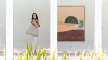 Load image into Gallery viewer, photo of young woman looking behind her while holding a stone coloured 100% linen Australian Made beach bag. Photo shot from outside window while she is standing in a windowed hallway
